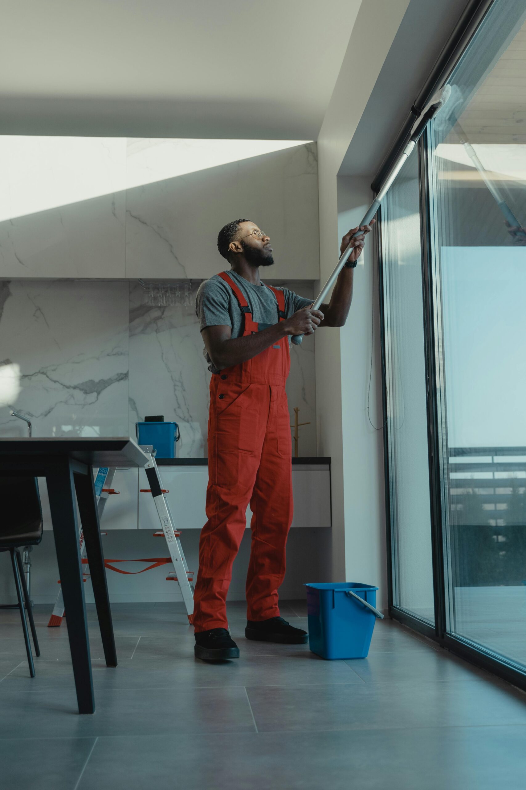 A man in coveralls cleans a large window in a stylish, modern home interior.