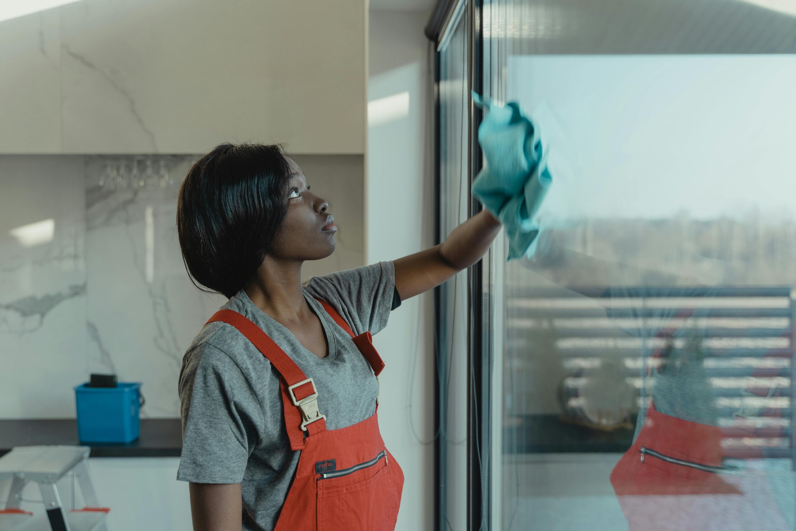 A woman in overalls cleans a glass window with a rag, emphasizing professional housekeeping.